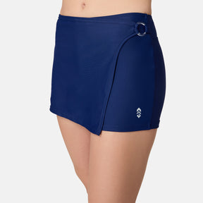 A person wears the Free Country Women's Fully Lined Swim Skort in navy, featuring a side ring detail and small white logo near the hem, photographed from waist to mid-thigh against a plain white background.