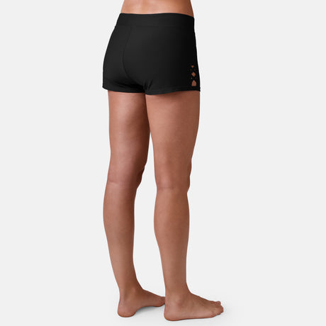A person stands barefoot facing away, modeling Free Country's Women's Laser Cut Boyshort in black, featuring small side crisscross cutouts and crafted from recycled materials, set against a plain light background.