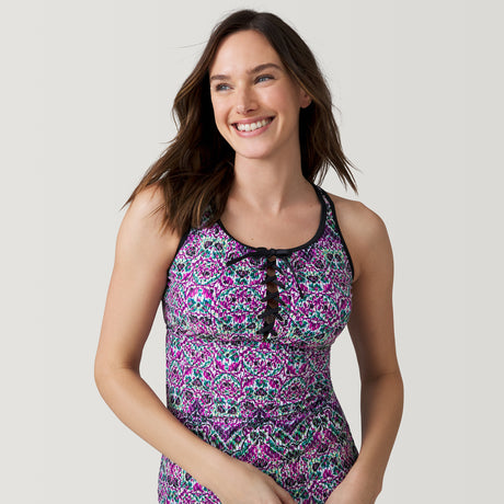 A woman with long brown hair smiles wearing the Women's Mosaic Mirage Lace Up Racerback Tankini Top by Clearance, featuring a pink, purple, black, and white mosaic print. She stands against a plain light gray background.