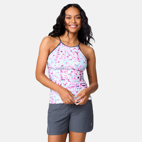 A woman with long dark hair smiles while wearing the Clearance Women's Mirage High Neck Macrame Back Tankini Top in a pink and blue pattern, paired with gray shorts, standing against a plain light background.