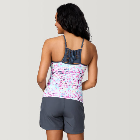 A woman with long dark hair, seen from behind, wears the Clearance Women's Mirage High Neck Macrame Back Tankini Top in a colorful abstract print and dark gray shorts, standing against a plain light background.