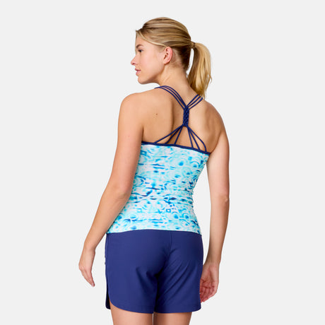 A woman with blonde hair in a ponytail is wearing the Clearance Women's Mirage Macrame Back Tankini Top, featuring a blue pattern and strappy back, paired with solid navy swim shorts, standing with her back to the camera against a white background.