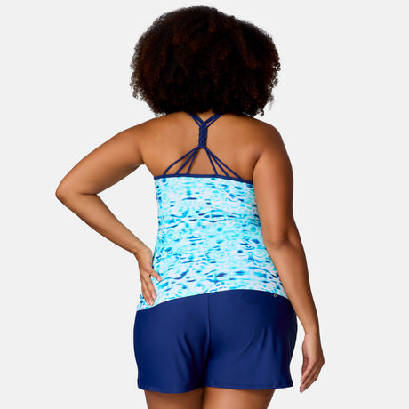 A woman with curly hair is seen from the back wearing the Clearance Women's Plus Size Mirage Macrame Back Tankini Top in blue and aqua, featuring crisscross straps, and matching navy swim shorts. One hand is on her hip.