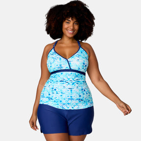 A woman with curly hair smiles while wearing the Women's Plus Size Mirage Macrame Back Tankini Top by Clearance and solid blue swim shorts, standing against a plain light background.