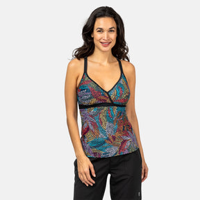 A woman with long dark hair wears the Clearance Women's Fresco Macrame Back Tankini Top, featuring a colorful design and UPF 50+ support, paired with black pants, standing and smiling against a plain light gray background.