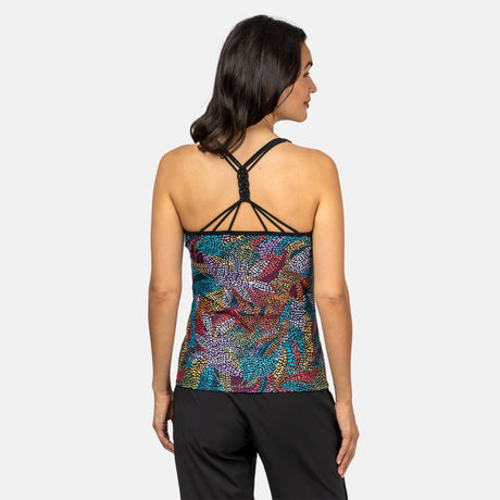 A woman with shoulder-length, wavy dark hair stands against a plain light gray background, wearing the Clearance Women's Fresco Macrame Back Tankini Top with colorful patterns, black crisscrossed straps, and black pants. The top offers a supportive fit.