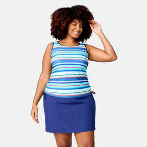 A smiling woman with curly hair wears the Women's Plus Size Marina Stripe Full Side Shirring Tankini Top by Clearance, paired with a blue skirt featuring front pockets, standing against a plain light background.