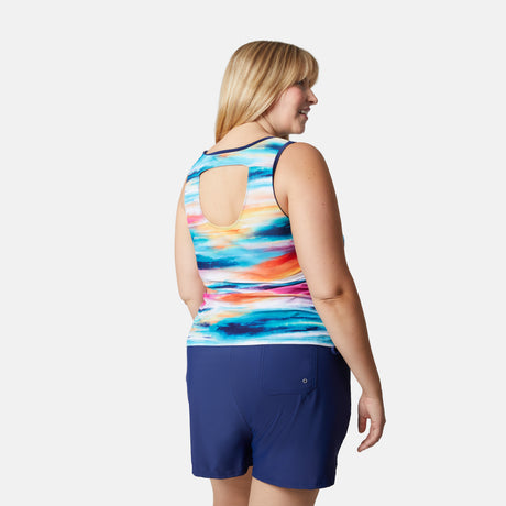 A blonde woman models the Free Country Women's Plus Size Horizon Full Side Shirring Tankini Top with an open back, paired with navy shorts, against a plain white background.