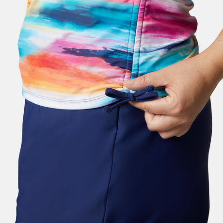 A person adjusts the side drawstring of a Free Country Women's Plus Size Horizon Full Side Shirring Tankini Top, featuring colorful fabric and paired with navy blue bottoms. The close-up captures their hand and the detail of the recycled material.