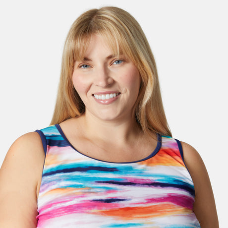 Smiling woman with long blonde hair and bangs wears the Free Country Women’s Plus Size Horizon Full Side Shirring Tankini Top, featuring a colorful striped, watercolor-like design, made from recycled polyester, against a plain white background.