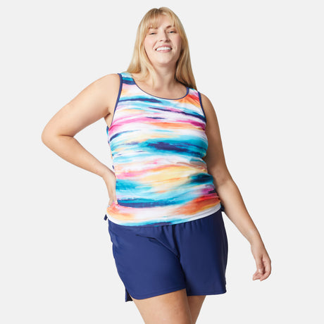 A smiling woman with long blonde hair wears the Free Country Women's Plus Size Horizon Full Side Shirring Tankini Top, featuring a colorful design, paired with navy blue shorts, standing confidently against a plain white background.