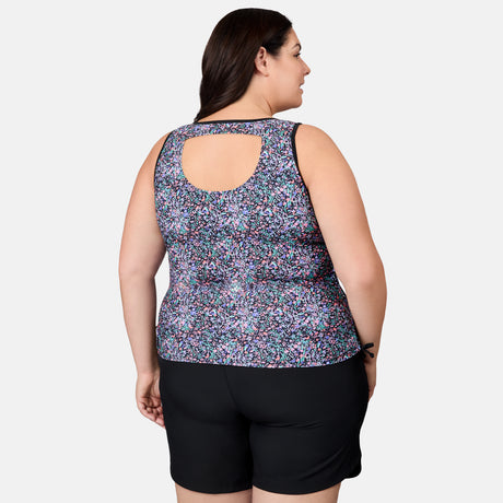 A woman is seen from the back in the Free Country Women's Plus Size Bursting Blooms Full Side Shirring Tankini Top, featuring a sleeveless, floral design with upper back cutout and quick-drying fabric, paired with black shorts against a white background.
