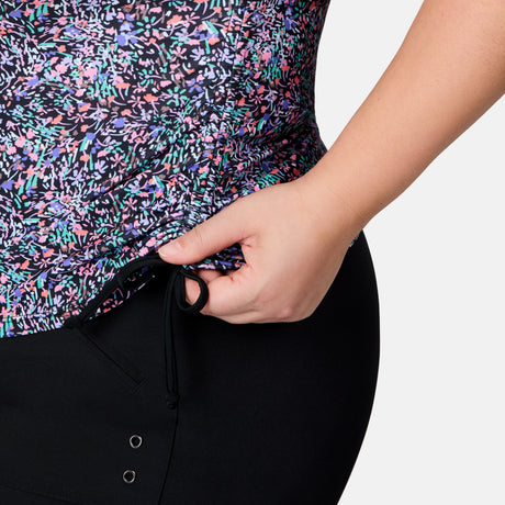 A person adjusts the drawstring on black pants, wearing the Free Country Women's Plus Size Bursting Blooms Full Side Shirring Tankini Top in a colorful floral print. The image is cropped to show only their lower torso and hand.