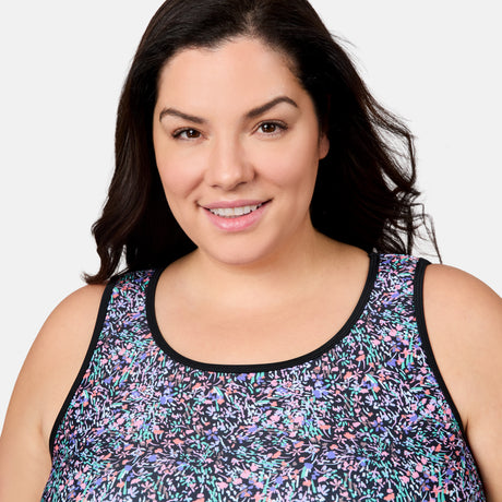 A woman with long dark hair smiles slightly at the camera, modeling the Free Country Women's Plus Size Bursting Blooms Full Side Shirring Tankini Top with a colorful floral pattern and black trim against a plain, light background.