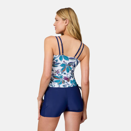 A woman with long blonde hair stands with her back to the camera, wearing the Free Country Women's Sunlit Floral Side Shirred V-Neck Tankini Top and navy swim shorts against a plain white background.
