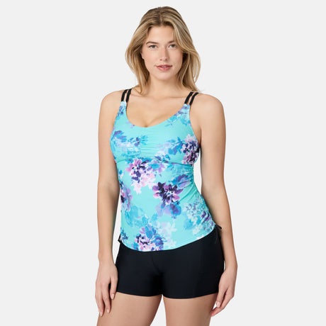 A woman wears the Free Country Women's Floral Zephyr Side Shirred V-Neck Tankini Top with adjustable double black straps and black swim shorts, offering UPF 50+ sun protection, against a plain white background, smiling at the camera.