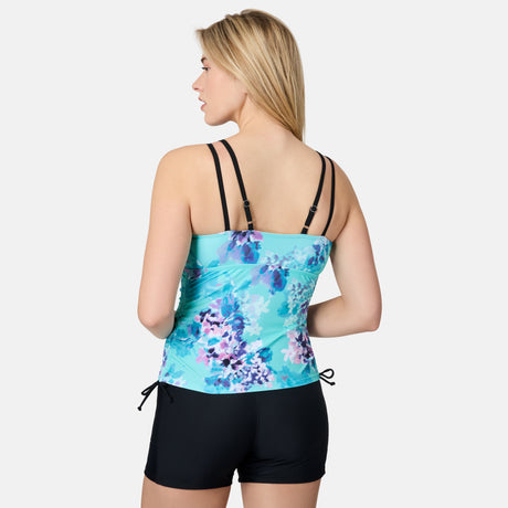A woman with long blonde hair faces away, wearing the Free Country Women's Floral Zephyr Side Shirred V-Neck Tankini Top with black adjustable straps and black swim shorts against a plain light gray background.