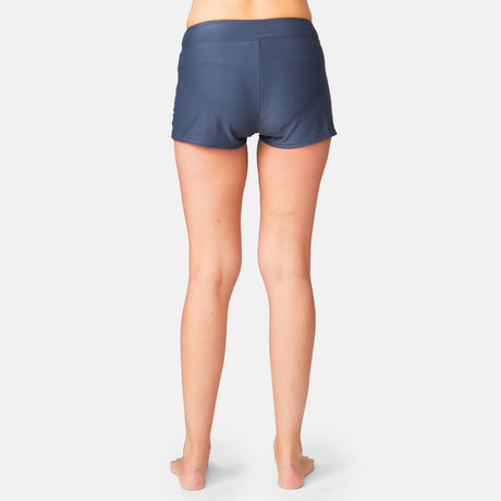 A woman stands barefoot on a white background, shown from the back, wearing Free Country Women's Laser Cut Boyshort swim bottoms in dark blue made from recycled materials.