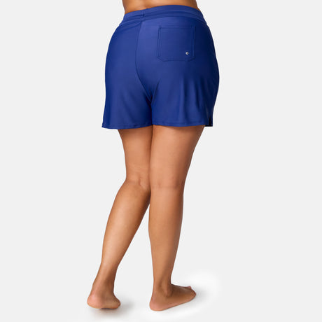 A woman stands barefoot, facing away, wearing Free Country Women's Plus Size Drawstring Swim Short in royal blue with a single back pocket and UPF 50+ protection, set against a plain light gray background.