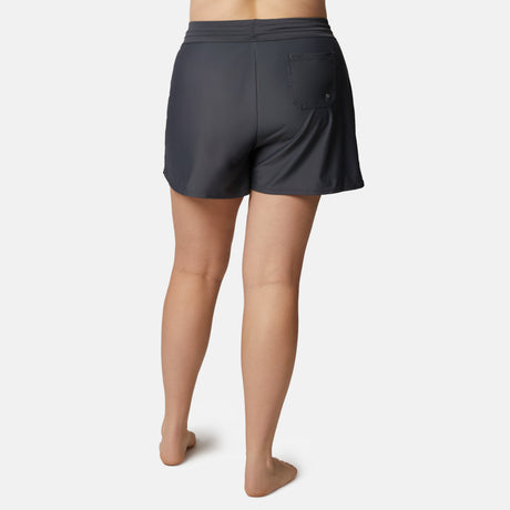 A woman is shown from the back, barefoot, wearing Free Country Women's Plus Size Drawstring Swim Short in dark gray with a single back pocket, against a plain white background.