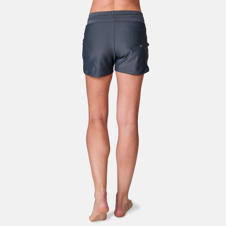A woman stands barefoot facing away, wearing Free Country Women's Drawstring Swim Short in dark grey with a back pocket and UPF 50+ protection, shown against a plain white background.