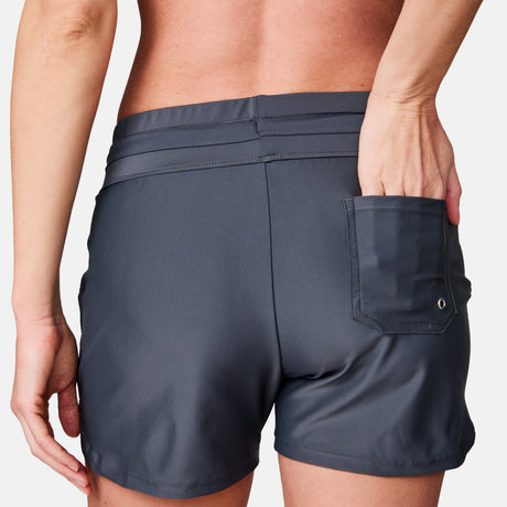 A woman wearing Free Country Women's Drawstring Swim Shorts in dark gray stands with her back to the camera, one hand partially tucked into the back pocket of the quick-dry shorts.
