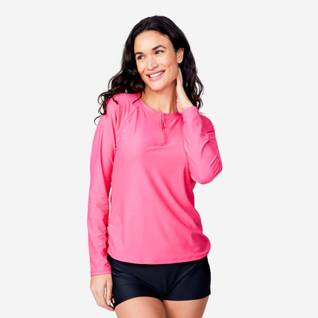A woman smiles with one hand on her neck, wearing the Clearance Women's Long Sleeve Zip Swim Shirt with Built-in Bra in bright pink and black shorts, standing against a plain white background.