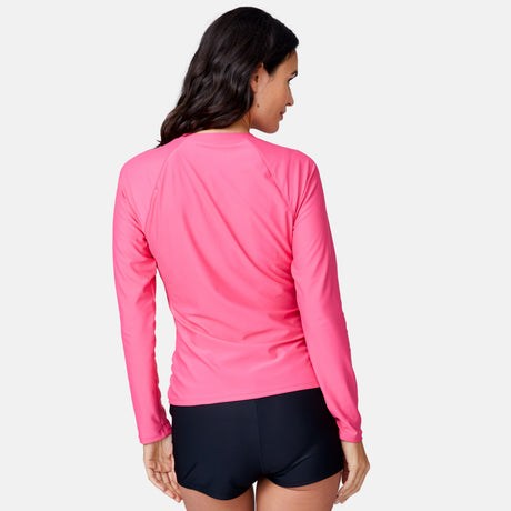 A woman with long dark hair wears the Clearance Women's Long Sleeve Zip Swim Shirt with Built-in Bra (bright pink) and black shorts, standing facing away from the camera against a plain light background.