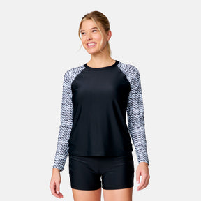 A woman smiles wearing the Clearance Women's Long Sleeve Swim Shirt with Built-in Bra and matching black swim shorts, featuring patterned sleeves, against a plain light background.