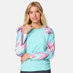 A woman with long blonde hair wears the Clearance Women's Long Sleeve Swim Shirt with Built-in Bra, UPF 50+, in light blue with colorful geometric sleeves, standing against a plain light gray background.