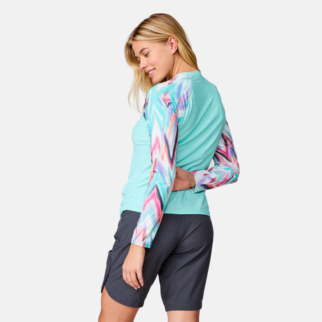 A woman stands sideways, smiling over her shoulder. She wears the Clearance Women's Long Sleeve Swim Shirt with Built-in Bra, featuring colorful patterned sleeves, and dark gray shorts against a plain light background.