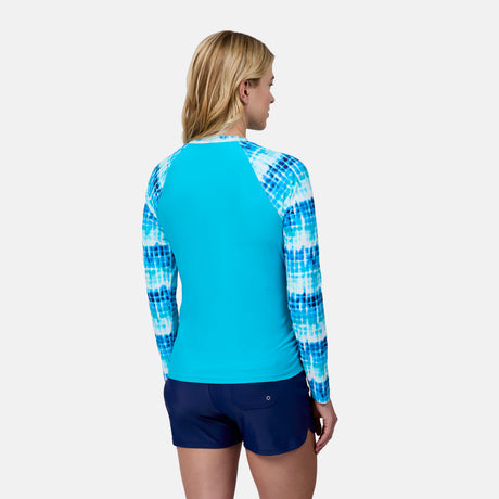 A woman with blonde hair, seen from the back, wears the Free Country Women's Long Sleeve Swim Shirt with Built-in Bra featuring blue and white tie-dye sleeves and navy swim shorts, standing against a plain light background.