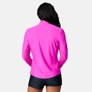 A woman with long dark hair faces away, wearing the Free Country Women's Sun Breeze 1/2 Zip Sunshirt in bright pink and black shorts against a plain white background.