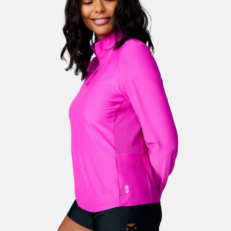 A woman smiles in the Free Country Women's Sun Breeze 1/2 Zip Sunshirt, a bright pink, moisture-wicking top made from recycled polyester and offering sun protection. She pairs it with black shorts against a plain white background.