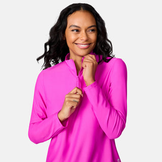 A woman with long dark wavy hair smiles as she zips up the bright pink Free Country Women's Sun Breeze 1/2 Zip Sunshirt against a plain white background.