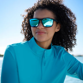 A woman with curly hair, wearing mirrored sunglasses and the Free Country Women's Sun Breeze 1/2 Zip Sunshirt, smiles outdoors against a backdrop of blue sky and water.