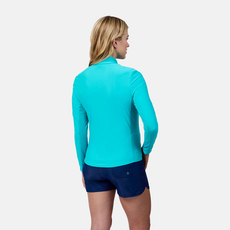 A woman with blonde hair stands facing away, wearing the Free Country Women's Sun Breeze 1/2 Zip Sunshirt in bright turquoise and navy shorts against a plain light background.