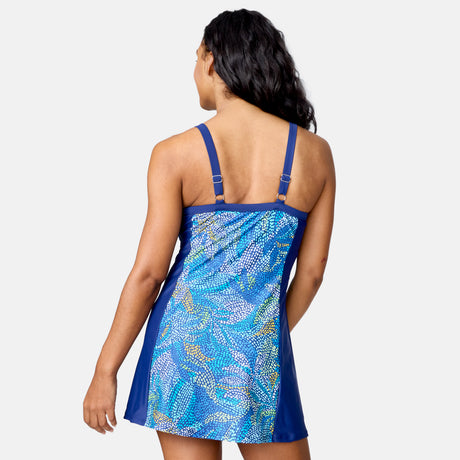 A woman with long, wavy hair faces away, modeling the Free Country Women's Swim Dress with Built-in Biker Short. The blue swim dress features adjustable straps and a colorful dotted back, blending comfort and style against a plain light background.
