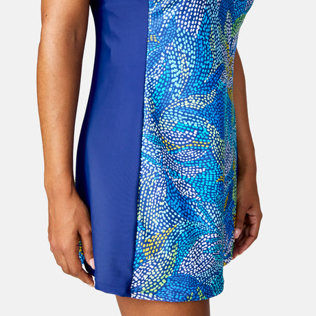 A woman wears the Free Country Women's Swim Dress with Built-in Biker Short, featuring a sleeveless design and blue multicolored abstract dotted accents. The photo shows only her torso and arms against a plain white background.