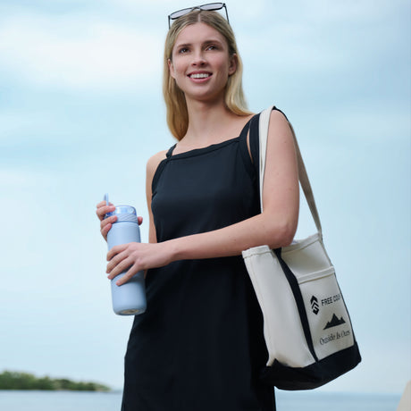 A woman in a black Free Country Women's Swim Dress with Built-in Biker Short stands outdoors, smiling, holding a reusable water bottle and a cream tote bag with "FREE COUNTRY" and "Outside is Ours." Sunglasses rest on her head.