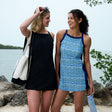 Two women stand by the water, smiling at each other. One wears the Free Country Women's Swim Dress with Built-in Biker Short, holding sunglasses and a tote; the other wears a blue-patterned quick-dry beach dress. Trees and water surround them.