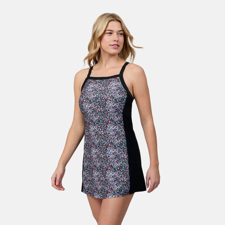 A woman models the Free Country Women's Swim Dress with Built-in Biker Short, featuring a sleeveless floral design and black side panels, as she stands against a plain light background looking left and smiling.
