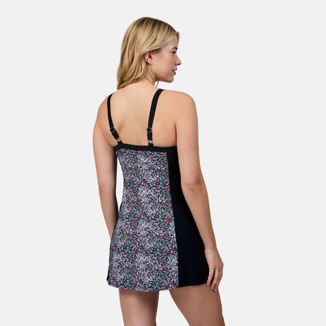 A blonde woman is seen from the back wearing the Free Country Women's Swim Dress with Built-in Biker Short, featuring UPF 50+, adjustable straps, a floral print center panel, solid black sides, and a built-in bra against a plain background.