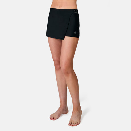 A person stands barefoot against a plain white background, wearing the Free Country Women's Fully Lined Swim Skort, featuring a wrap-around design, built-in swim bottom, and a small logo on the left thigh.