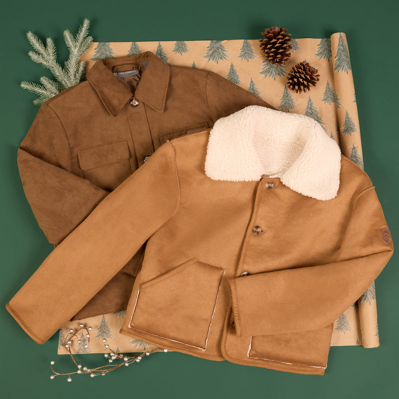 Two brown suede-like jackets arranged on a patterned wrapping paper with pine branches and pinecones, set against a dark green background.