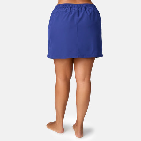 A woman stands barefoot, back to camera, wearing the Free Country Women's Plus Size Beach and Beyond Skort in royal blue—knee-length with an elastic waistband and four-way stretch—against a plain white background.