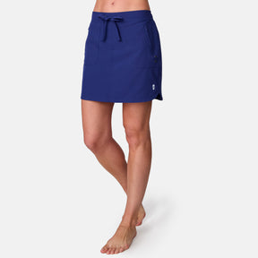A woman models the Free Country Women's Beach and Beyond Skort, featuring front pockets, a drawstring waist, and four-way stretch fabric in blue, standing barefoot against a plain white background.