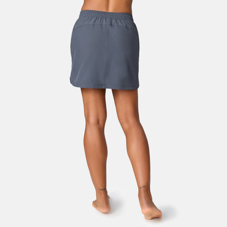 A person stands barefoot facing away, wearing the Free Country Women's Beach and Beyond Skort, which features an elastic waistband and four-way stretch. The background is plain white.
