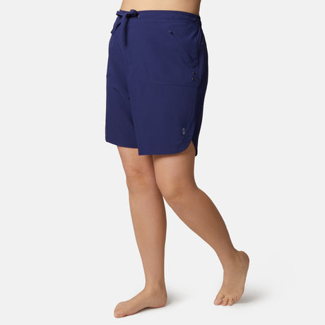 A woman stands barefoot in Free Country Women's Plus Size Bermuda Board Short II, featuring quick-drying fabric, front pockets, and a drawstring waist, against a plain white background.