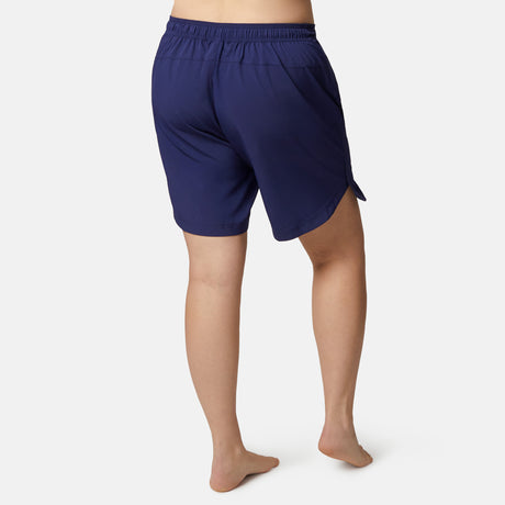 A woman stands barefoot, facing away, in Free Country’s Women's Plus Size Bermuda Board Short II—navy blue with an elastic waistband and side slits. The quick-dry fabric is ideal for active days, shown on a plain white background.